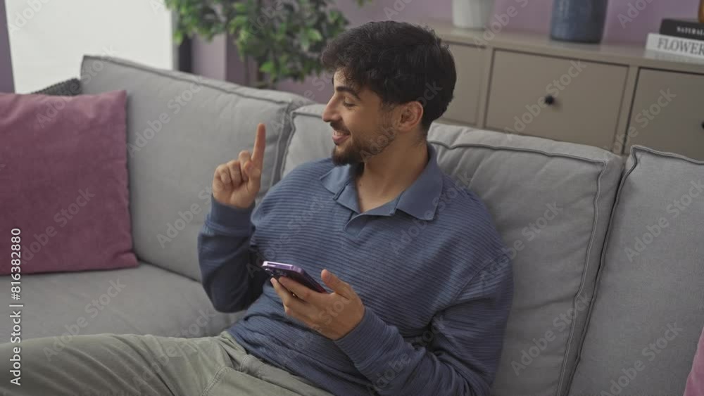 Young, happy arab man sitting at home on sofa, smiling and pointing to side with one hand while using smartphone Young, happy arab man sitting at home on sofa, smiling and pointing to side with one hand while using smartphone