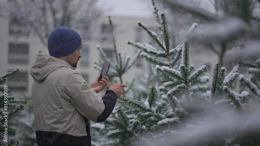Man video blogger shoots video on his phone for social networks and subscribers on video services about the choice of a Christmas tree at the street before Christmas market in the snowy weather. Man video blogger shoots video on his phone for social networks and subscribers on video services about the choice of a Christmas tree at the street before Christmas market in the snowy weather.