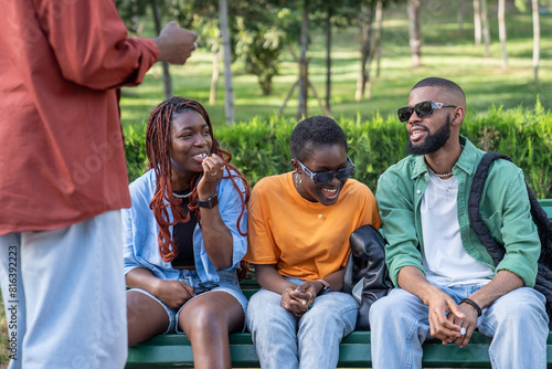 Happy carefree four african american friends girls and guys laughs have fun together sitting on park bench. Serene joyful black students smiling chats sharing good mood and gossip stories rest outdoor