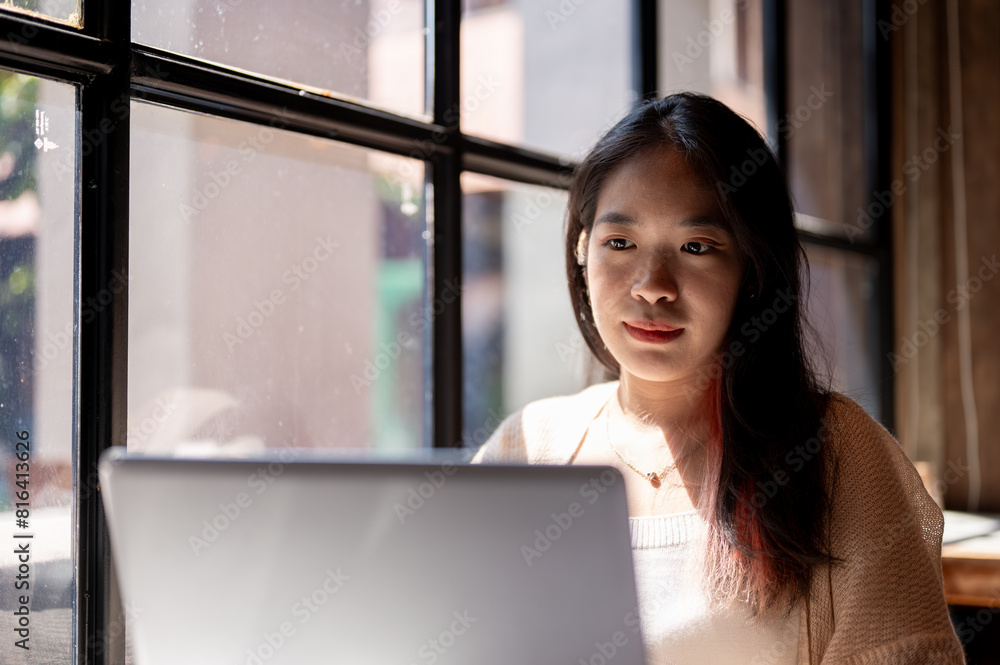 Young focused Asian woman working remotely at a coffee shop, working on her laptop computer. Young focused Asian woman working remotely at a coffee shop, working on her laptop computer.