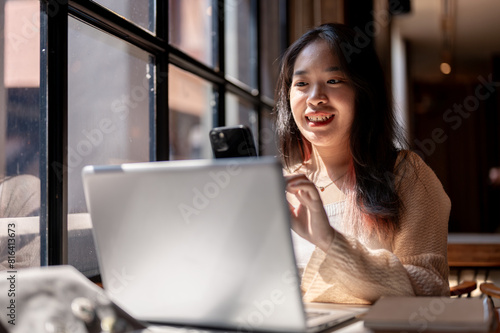 Young cheerful Asian woman using her smartphone while sitting in a coffee shop with her laptop.