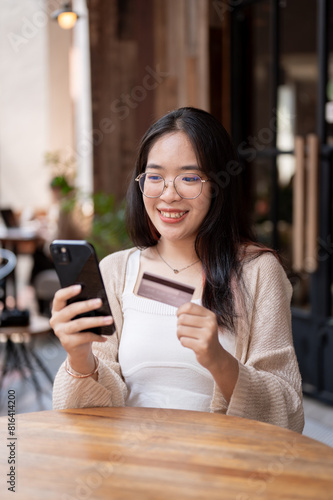 A happy Asian woman sits at an outdoor table of a cafe, holding her credit card and smartphone.