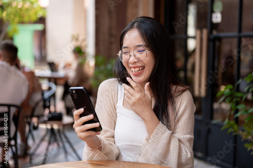 A cheerful Asian woman enjoys talking on a video call while sitting at an outdoor table of a cafe.