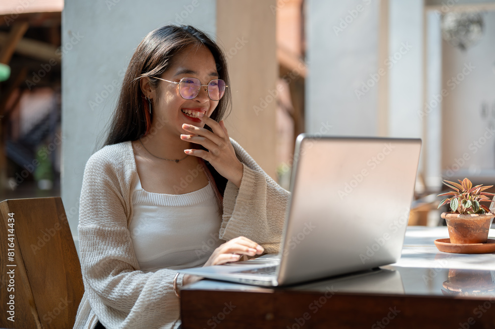A cheerful Asian woman is laughing while enjoying talking on a video call on her laptop at a cafe. A cheerful Asian woman is laughing while enjoying talking on a video call on her laptop at a cafe.