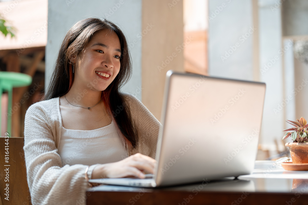 A beautiful, happy Asian woman is sitting outdoors, working remotely at a cafe, using her laptop. A beautiful, happy Asian woman is sitting outdoors, working remotely at a cafe, using her laptop.
