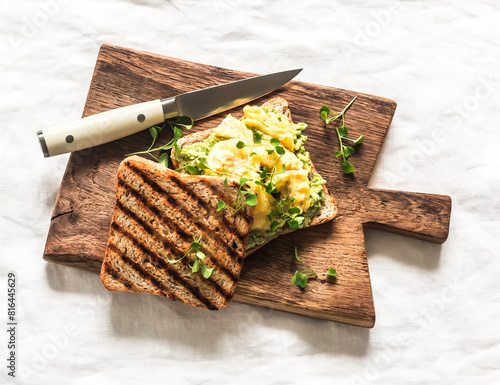 Delicious breakfast, brunch, snack - wholegrain toast with avocado, scrambled eggs and microgreens on a wooden chopping board on a light background, top view