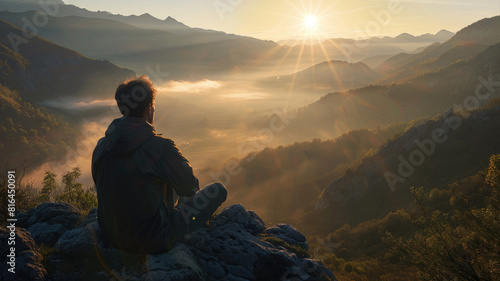 A man sitting from the back overlooks a stunning valley with sunlight breaking through the mist