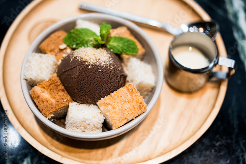 A delectable chocolate ice-cream served with sliced bread and a shot of sweetened condensed milk on a wooden plate is placed on a wooden table at a cafe. Dessert food menu photo, selective focus.