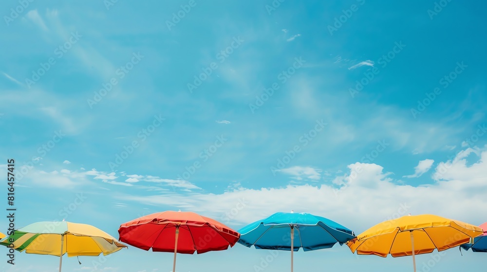 Six colorful beach umbrellas are arranged in a row against a blue sky with white clouds. The umbrellas are red, blue, green yellow and orange. Six colorful beach umbrellas are arranged in a row against a blue sky with white clouds. The umbrellas are red, blue, green yellow and orange.