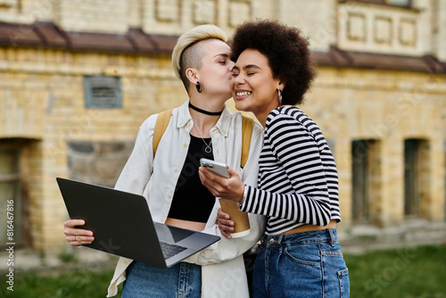 Two young women in casual attire stand in front of a building, working on a laptop. photo