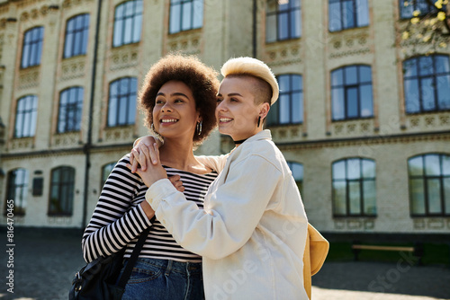 Two young women in casual attire embrace in front of a university building.