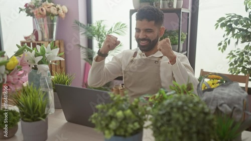 Hispanic man with beard celebrating success on laptop in a flower shop indoor setting. photo