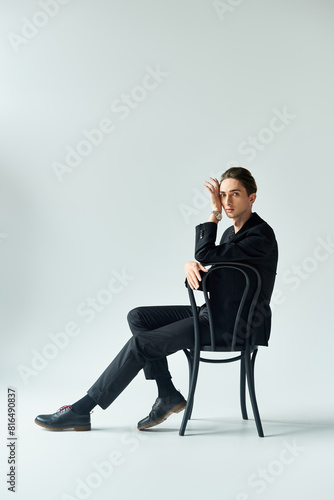 A stylish young man in a suit sits on a chair, exuding confidence and contemplation in a studio with a grey background.