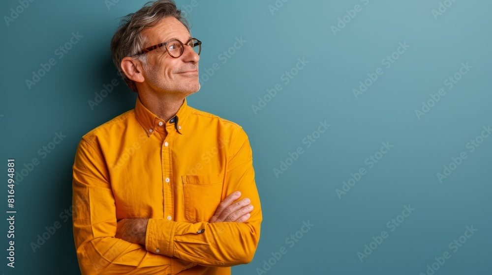 Fototapeta premium A man in a yellow shirt and glasses stands with crossed arms before a blue backdrop