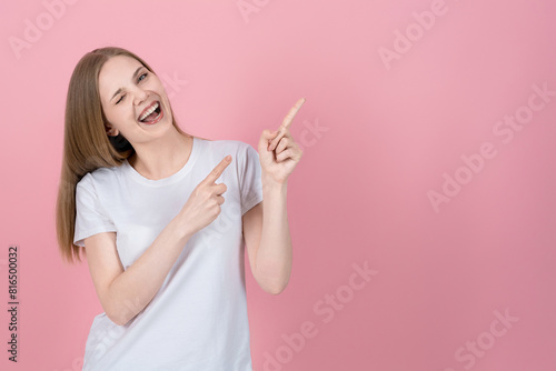 Attractive caucasian young blonde woman in casual white t-shirt smiling, winking and pointing fingers at empty copy space for advertising on pink studio background. Sale, advice.