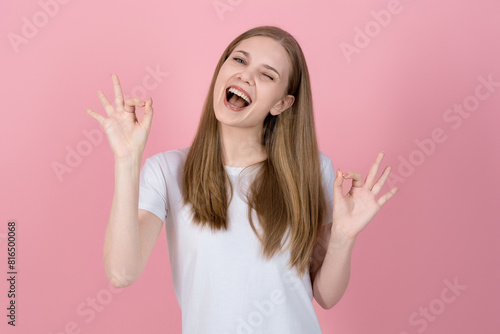Attractive caucasian young blonde woman in casual white t-shirt smiling friendly, winking and showing ok gesture isolated on pink studio background. Good, Success, Approved.