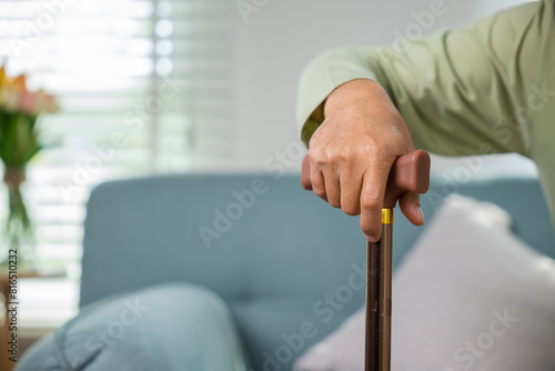 Asian senior old man with eyeglasses type to standing up from sofa with walking cane stick to walk at home, Elderly suffering from knee pain ache holding handle of cane, retirement medical healthcare