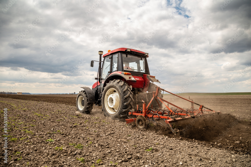 Obraz premium Powerful red tractor tills the soil on a vast farm with a dramatic cloudy sky overhead