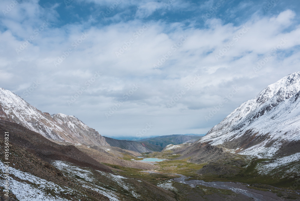 Dramatic landscape with river meanders and mountain lake in green stone valley among snow covered hills in rainy cnangeable weather. Freshly fallen snow in high mountains under clouds in blue sky.