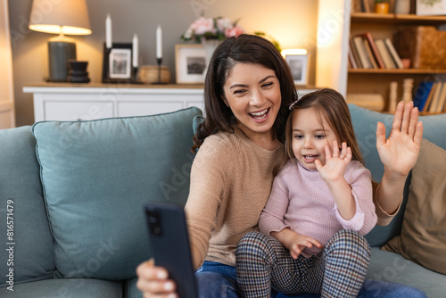 Happy mother and kid daughter waving hands looking at phone camera for video call, smiling mom and child girl having fun greeting online by phone webcam making videocall with grandparents.