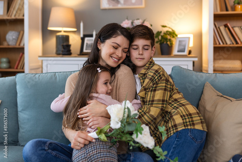 Young mother with a bouquet of roses laughs, hugging her son, and cheerful girl with a flowers congratulates mom during holiday celebration at home