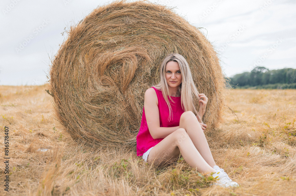 many bale of straw dried grass in a field, food for animals. big rolls and blonde girl relaxing on it.women in short pants posing in meadow. sunny summer day. many bale of straw dried grass in a field, food for animals. big rolls and blonde girl relaxing on it.women in short pants posing in meadow. sunny summer day.