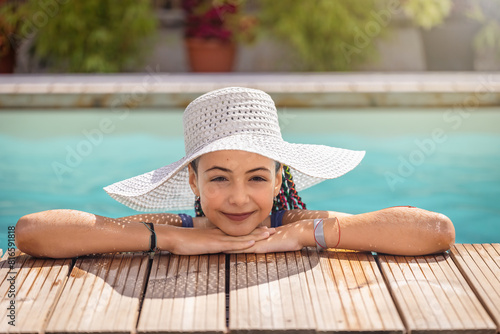 Little smiling girl with white straw hat is leaning on the edge of the pool looking at the camera.  Horizontally. 
