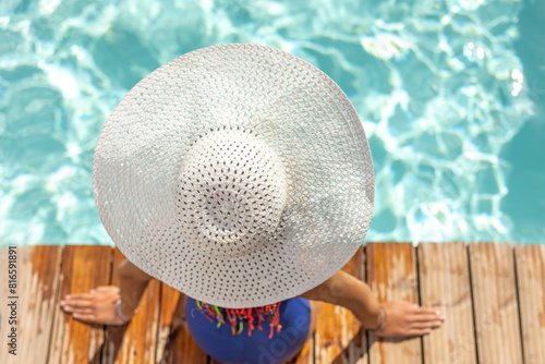 Top view of little girl with white straw hat sitting on edge of pool with feet in water. Horizontally. 