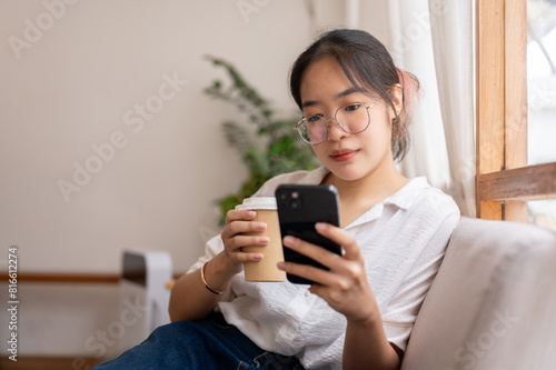 A woman reading online blog on her smartphone while sipping coffee on the sofa in the living room.