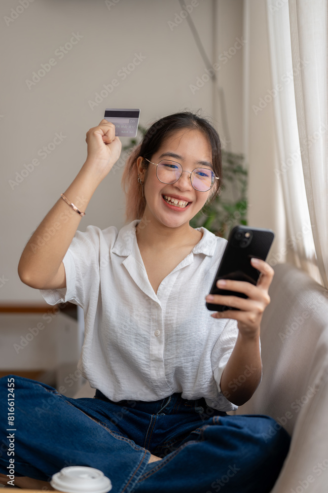 A cheerful Asian woman holding a credit card and a smartphone, enjoying shopping online at home. A cheerful Asian woman holding a credit card and a smartphone, enjoying shopping online at home.