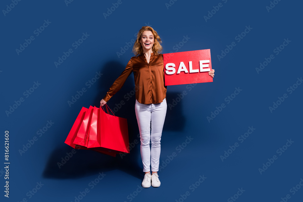 Full length photo of friendly nice woman dressed silk shirt hold shopping bags red sale placard isolated on dark blue color background Full length photo of friendly nice woman dressed silk shirt hold shopping bags red sale placard isolated on dark blue color background