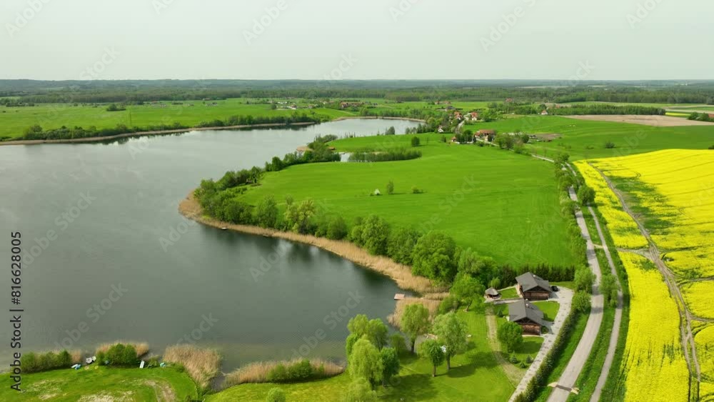 Aerial view of a lake surrounded by green fields, small houses, and a rapeseed field, under a clear sky Aerial view of a lake surrounded by green fields, small houses, and a rapeseed field, under a clear sky