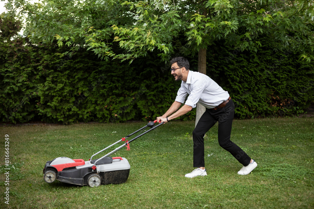 Cheerful businessman mowing the lawn with a lawnmower in the backyard. Leisure and household concept Cheerful businessman mowing the lawn with a lawnmower in the backyard. Leisure and household concept