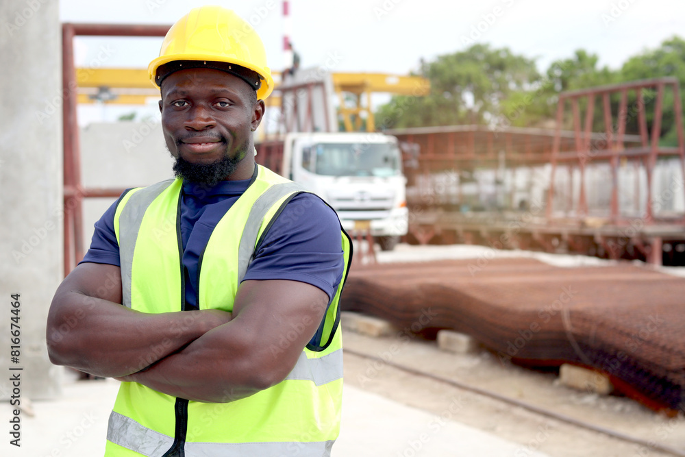 Portrait of happy smiling African engineer worker with safety vest and helmet standing with arms crossed at construction site. Young architect working at ground level of building site workplace. Portrait of happy smiling African engineer worker with safety vest and helmet standing with arms crossed at construction site. Young architect working at ground level of building site workplace.