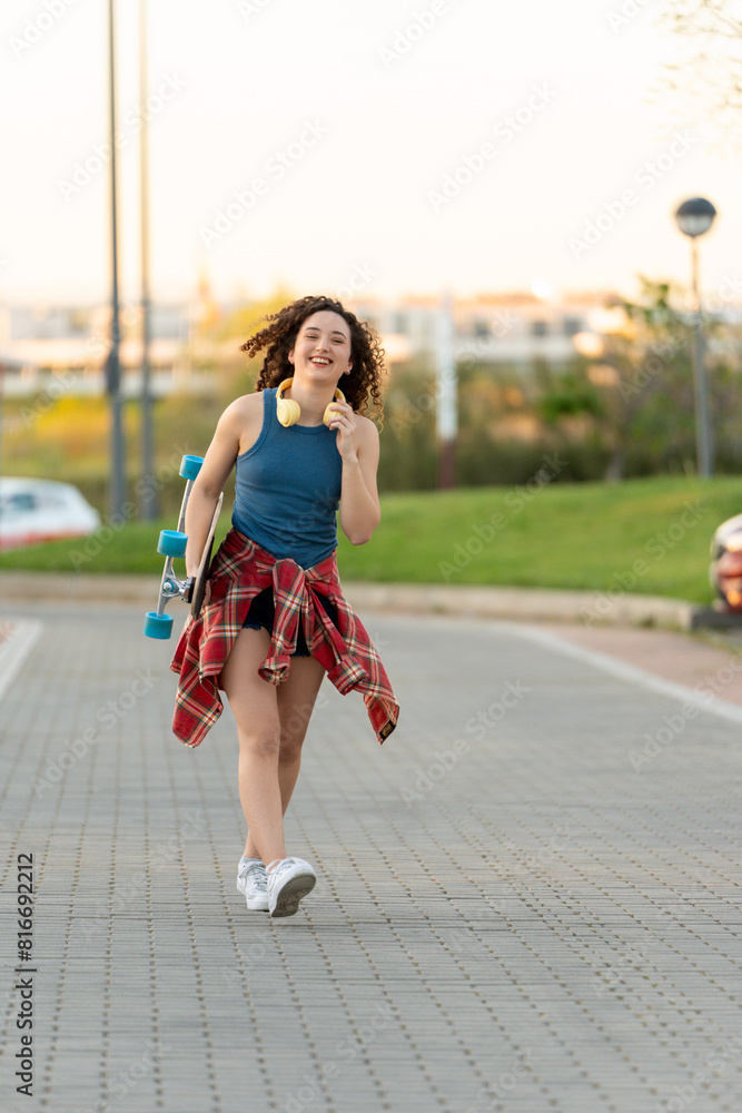 Cheerful curly-haired woman carrying a skateboard and wearing headphones, strolling in a park, with a blue tank top and plaid shirt at sunset. Cheerful curly-haired woman carrying a skateboard and wearing headphones, strolling in a park, with a blue tank top and plaid shirt at sunset.