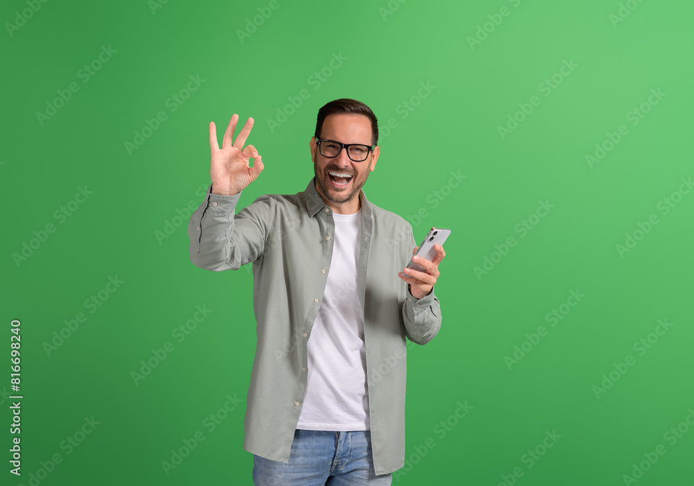 Young man laughing cheerfully and showing OK gesture while using mobile phone on green background Young man laughing cheerfully and showing OK gesture while using mobile phone on green background