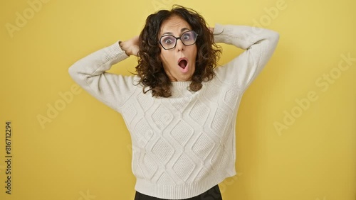 Shocked! crazy-scared middle-aged hispanic woman with glasses, standing hands-on-head, mouth open wide in surprise against a yellow cut-out background photo