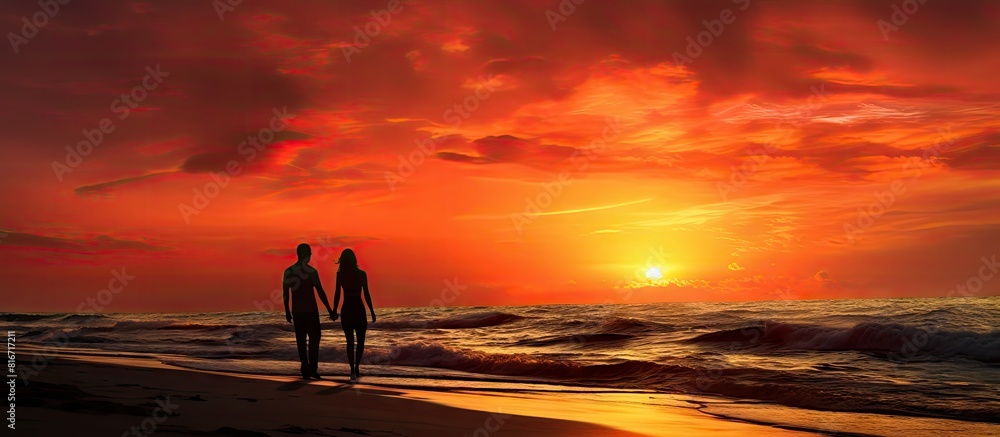 A stunning sunset by the sea with a couple captivated by the mesmerizing sight The red yellow sky creates a breathtaking backdrop as silhouettes of people add to the enchantment of this evening at th
