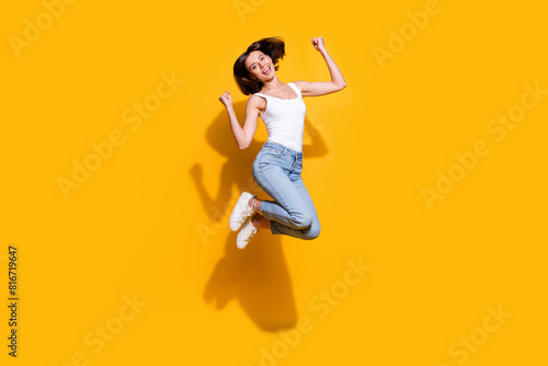 Full body photo of attractive young girl raise fists jumping excited wear trendy white outfit isolated on yellow color background