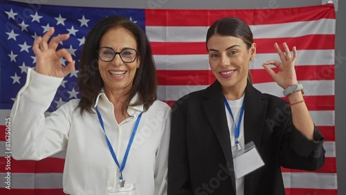 Two professional women with id badges standing before an american flag in an office, making positive gestures. photo