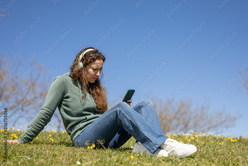 Beautifu l young woman enjoying music with headphones outdoors in a lush green park. Woman stops listening to the podcast to answer messages on her phone. Beautifu l young woman enjoying music with headphones outdoors in a lush green park. Woman stops listening to the podcast to answer messages on her phone.