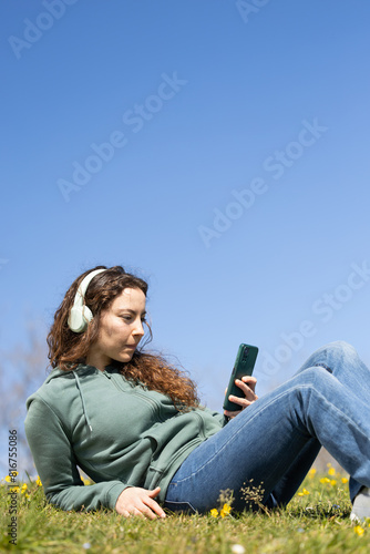 Beautifu l young woman enjoying music with headphones outdoors in a lush green park. Woman stops listening to the podcast to answer messages on her phone.