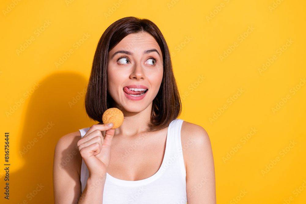 Photo portrait of attractive young girl hold cooky lick lips look empty space wear trendy white outfit isolated on yellow color background Photo portrait of attractive young girl hold cooky lick lips look empty space wear trendy white outfit isolated on yellow color background