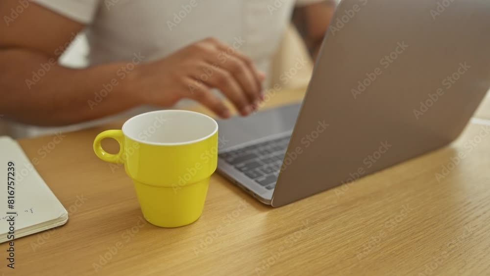 Adult black man using laptop in a modern living room with a yellow mug and notebook on the wooden table Adult black man using laptop in a modern living room with a yellow mug and notebook on the wooden table