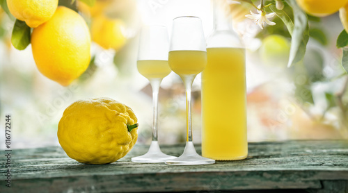 Bottle limoncello and two glasses standing on weathered wooden table. Background with lemon trees in backlight. Close-up.
