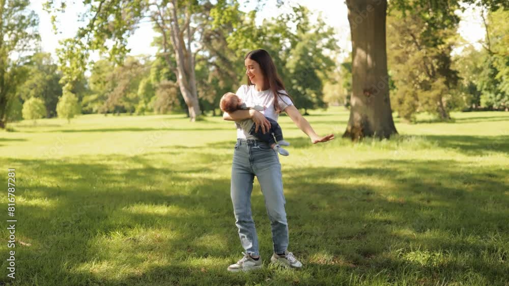 A young mother holds a baby in her arms, spins with him in the park during a walk in the summer in sunny warm weather A young mother holds a baby in her arms, spins with him in the park during a walk in the summer in sunny warm weather