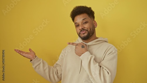 Gob-smacked african american guy in a hoodie, cheerfully pointing and presenting with hand, wearing a radiant smile on isolated yellow background photo