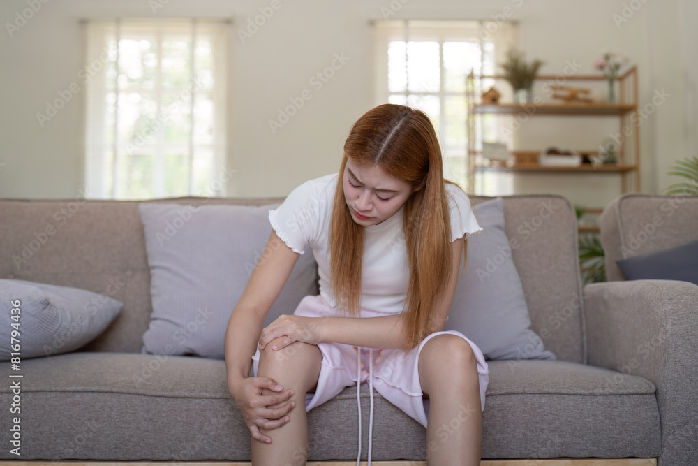 Young Asian woman experiencing knee pain while sitting on a sofa at home. Concept of health issues and discomfort Young Asian woman experiencing knee pain while sitting on a sofa at home. Concept of health issues and discomfort