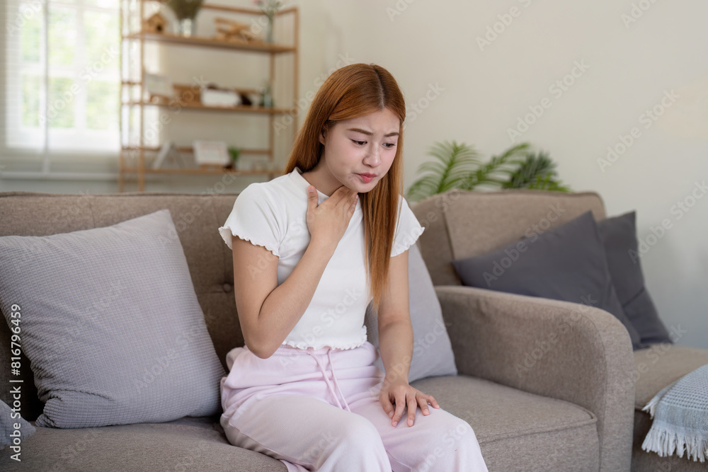 Young Asian woman holding her throat while sitting on a sofa. Concept of throat pain and health issues Young Asian woman holding her throat while sitting on a sofa. Concept of throat pain and health issues