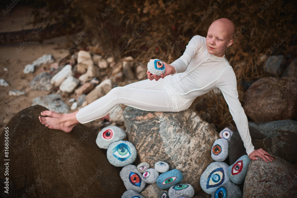 Naklejka premium Young hairless girl with alopecia in white futuristic costume pensively examines stone with eye at surreal landscape, performance symbolizes introspection and reevaluation of individuality